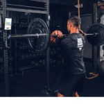 A man in a black shirt and shorts performs a front squat with a loaded barbell in a gym, as the Perch Camera captures his movement. Weight plates and gym equipment are visible in the background.