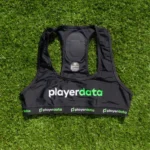 A PlayerData GPS black sports vest with "playerdata" in white and green text lies on green grass. It features an elastic band with playerdata branding and a built-in sensor pouch.