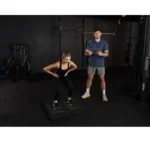 A woman performs a squat on exercise mats in a gym while a man with a clipboard observes her using VALD ForceDecks, with various gym equipment visible in the background.