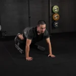 A man performs a bodyweight exercise on a flat bench, using the VALD NordBoard placed on a black rubber mat, with colorful weight plates visible on the wall behind him.