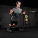 A man in athletic wear uses the VALD NordBoard, kneeling on its padded bench and leaning forward with hands at shoulder level, performing a Nordic hamstring exercise in a gym with weight plates on the wall behind him.