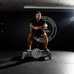 A focused man in athletic wear sits on a kBox Advanced System in a gym with a dark wall and large circular logo behind him, facing forward with hands on his knees.
