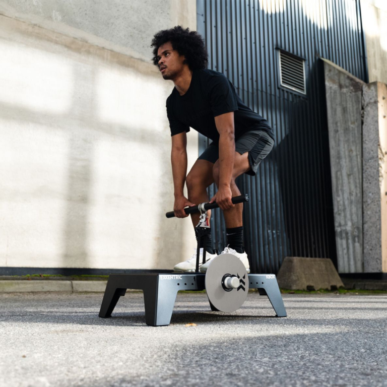 Wearing athletic gear, a person gets ready to lift with the kBox Core System on an outdoor platform, with a corrugated metal wall in the background.