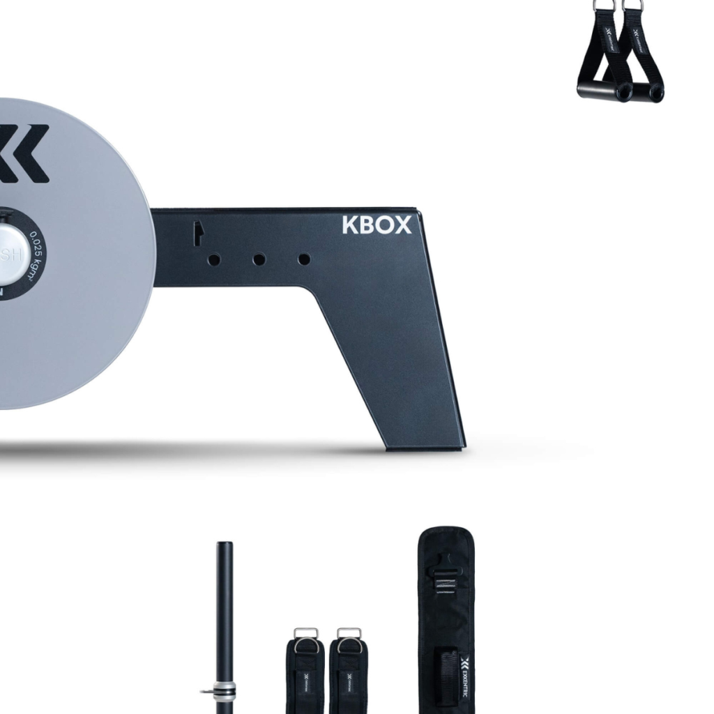 Partial view of the kBox Core System flywheel trainer with black and silver fitness accessories—straps, a bar, and a carrying case—arranged on a white background.