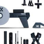 The kBox Ultimate System flywheel training device is shown with black and gray fitness accessories—straps, bars, and attachments—arranged around it on a white background.