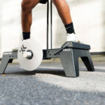 A person stands on a black kBox Advanced System with resistance bands and a flywheel, preparing to work out; only their legs and feet in sneakers are visible.